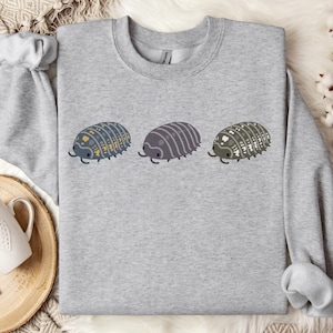 Könnte beinhalten: Ein hellgraues Sweatshirt mit Rundhalsausschnitt und einem Grafikdruck von drei Asseln in einer horizontalen Reihe. Die erste Assel ist blaugrau mit gelben Punkten, die zweite ist einfarbig lilagrau und die dritte ist dunkelgrau mit weißen Punkten.