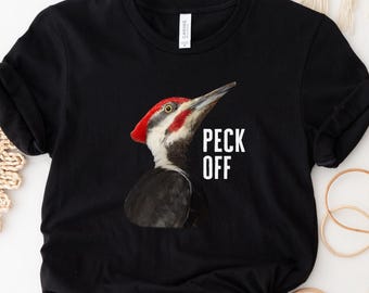 May include: Black t-shirt featuring a graphic of a Pileated Woodpecker with a red crest and the text "PECK OFF" in white. The shirt is made of a soft material and has rolled-up sleeves. A black circle with the word "BLACK" is in the lower left corner.
