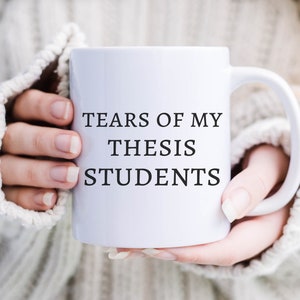 May include: White ceramic mug with black text that reads "Tears of my thesis students".