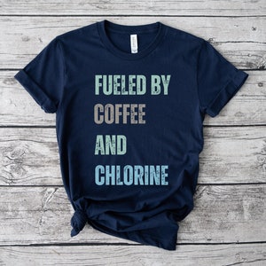 Puede incluir: Una camiseta azul marino con el texto "Fueled by Coffee and Chlorine" en una fuente desgastada. El texto está en tonos de verde, marrón y azul.