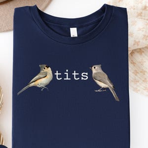 Puede incluir: Camiseta azul marino con la palabra "tits" en blanco entre dos ilustraciones de p&aacute;jaros. Los p&aacute;jaros son en tonos grises, marrones y blancos. La camiseta es de color azul marino liso.