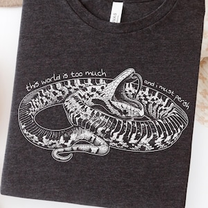 May include: A dark grey heather t-shirt featuring a white line art illustration of a coiled snake with its mouth open. The text "this world is too much" is printed above the snake, and "and i must perish" is printed to the right of the snake.
