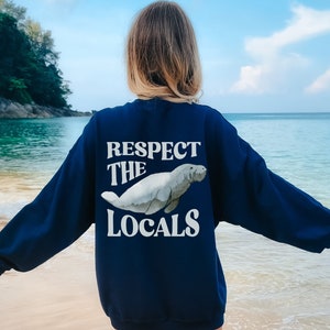 May include: Navy blue sweatshirt with the text "RESPECT THE LOCALS" and a watercolor illustration of a manatee. The sweatshirt is a long-sleeved crew neck style.
