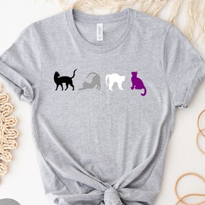 May include: A gray t-shirt with a row of four cats in black, gray, white, and purple. The cats are arranged in a line, with the black cat on the left and the purple cat on the right. The shirt is made of a soft, comfortable material and is perfect for everyday wear.