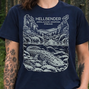 May include: A navy blue short-sleeve t-shirt featuring a light-coloured graphic print of a hellbender salamander resting on rocks in a stream, with a mountain landscape background and the text HELLBENDER APPALACHAIN MOUNTAIN STREAMS.