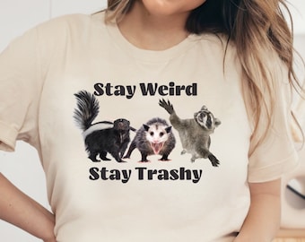 May include: A beige t-shirt with the text "Stay Weird Stay Trashy" printed on it. The text is in black and there are three animals printed below the text: a skunk, a possum, and a raccoon.