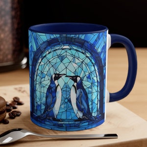 May include: A blue and white ceramic coffee mug with a stained glass design of two penguins looking at each other. The mug has a blue rim and handle.