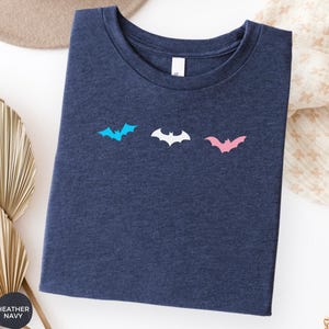 May include: A heather navy blue crew neck t-shirt featuring three small bat silhouettes printed in a horizontal row across the chest. The left bat is light blue, the center bat is white, and the right bat is light pink in color.