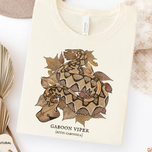 Può includere: T-shirt color crema con un'illustrazione dettagliata di una vipera del Gabon (Bitis gabonica) tra foglie autunnali. Il testo "Gaboon Viper (Bitis gabonica)" è stampato sotto. La maglietta è piegata.