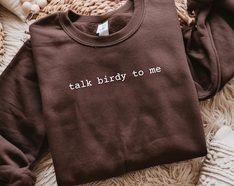 Puede incluir: Una sudadera marrón con la frase "talk birdy to me" impresa en blanco. La sudadera tiene cuello redondo y mangas largas con puños. El texto está en una fuente sencilla. La sudadera está doblada sobre una superficie tejida.