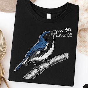 Puede incluir: Una camiseta negra con una ilustración gráfica de un pájaro azul y negro posado en una rama. El pájaro está representado con el pico abierto y el texto "I'M SO LA-ZEE" está impreso en letras blancas junto a la cabeza del pájaro.