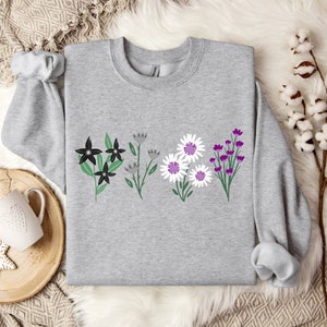 May include: A light gray sweatshirt featuring a floral design. The design includes black, gray, white, and purple flowers with green leaves. The sweatshirt has a crew neck and long sleeves.