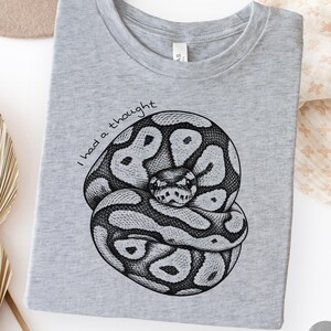 May include: A light grey t-shirt featuring a black ink-style illustration of a coiled ball python. The snake is positioned in the center with the handwritten text "I had a thought" arched above it in a simple, dark, sans-serif font style.