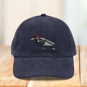Puede incluir: Gorra de béisbol de pana azul marino con un diseño de pájaro carpintero bordado y el texto "I WANT TO BELIEVE". El pájaro carpintero tiene la cabeza roja, el cuerpo negro y detalles blancos. La gorra tiene una visera curva.