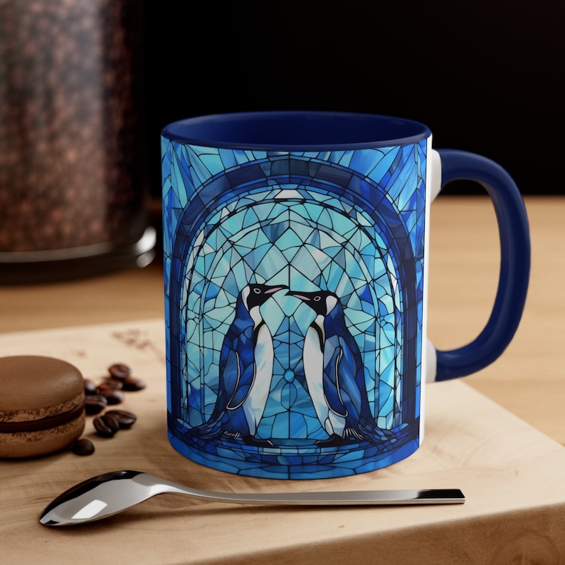 K&ouml;nnte beinhalten: Eine blaue und wei&szlig;e Keramik-Kaffeetasse mit einem Buntglas-Design von zwei Pinguinen, die sich ansehen. Die Tasse hat einen blauen Rand und einen blauen Griff.