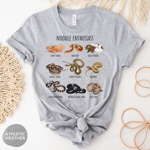 Puede incluir: Camiseta gris jaspeado con la frase "NOODLE ENTHUSIAST" y dibujos de serpientes como la serpiente del ma&iacute;z y la pit&oacute;n bola. La camiseta est&aacute; anudada a la cintura.