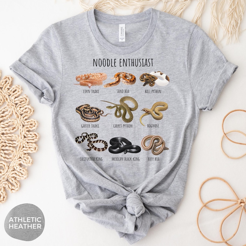 Puede incluir: Camiseta gris jaspeado con la frase "NOODLE ENTHUSIAST" y dibujos de serpientes como la serpiente del ma&iacute;z y la pit&oacute;n bola. La camiseta est&aacute; anudada a la cintura.