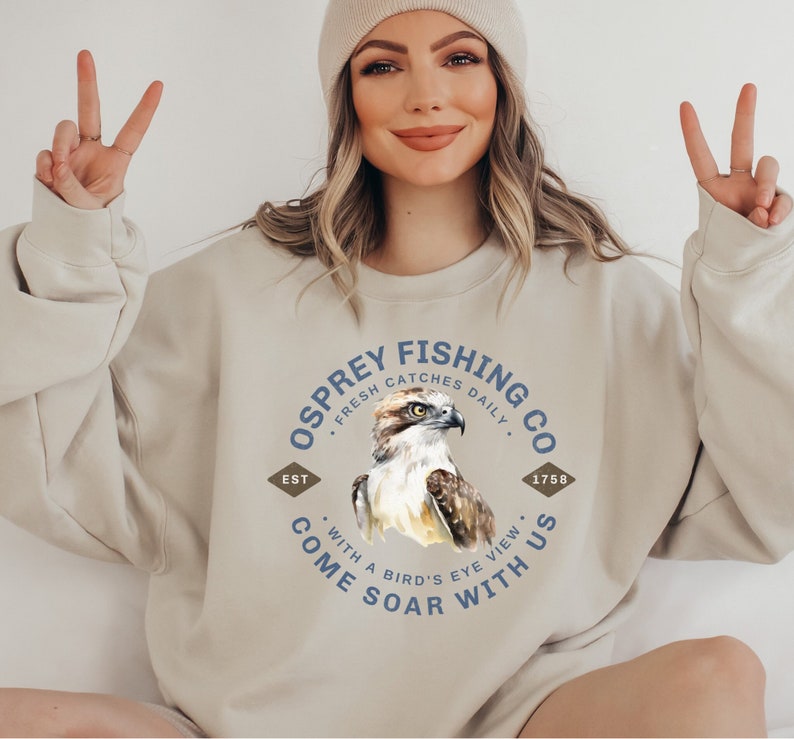 May include: A beige sweatshirt with a circular graphic design featuring an osprey bird, the text "Osprey Fishing Co." and the words "Fresh Catches Daily", "Est", "1758", "With a Bird's Eye View", "Come Soar With Us".