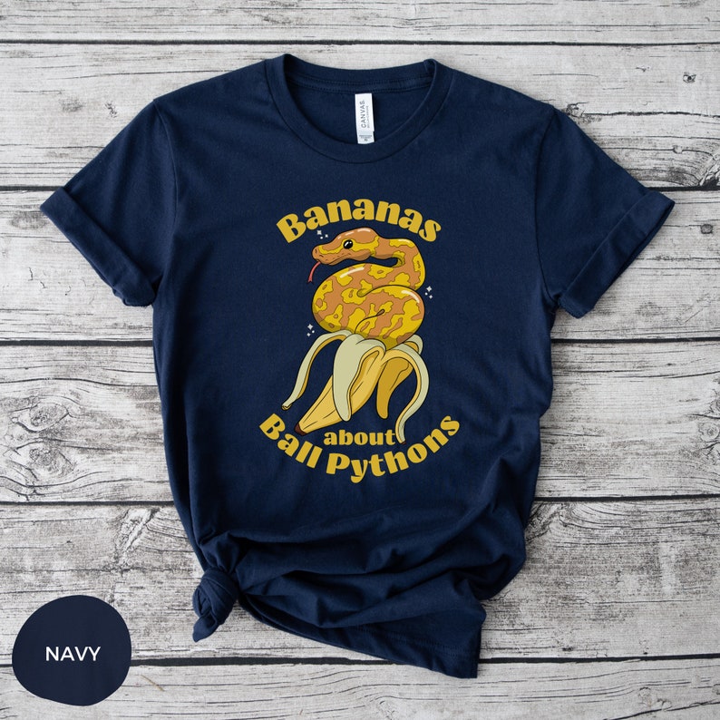 Op de afbeelding: Een marineblauw t-shirt met een afbeelding van een gele banaan met een gele en bruine ball python eromheen gewikkeld. De tekst "Bananas about Ball Pythons" staat op het shirt gedrukt.