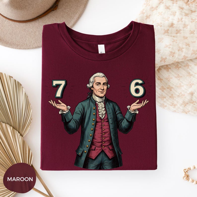 May include: Maroon t-shirt featuring a cartoon illustration of a man in a historical outfit, holding the numbers 7 and 6. The man is wearing a green jacket, red vest, and white ruffled shirt. The numbers are in a glowing font.