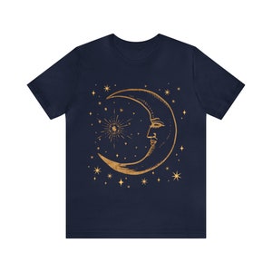 Pode incluir: Uma camiseta azul marinho com um design de lua crescente dourada e estrelas. A lua tem um rosto e &eacute; cercada por pequenas estrelas.