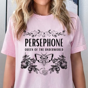 May include: Pink t-shirt with a black graphic design featuring the text "PERSEPHONE QUEEN OF THE UNDERWORLD" with a floral design and a moth with a skull.