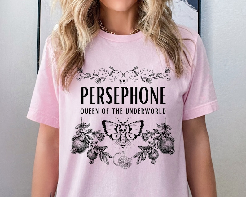 May include: Pink t-shirt with a black graphic design featuring the text "PERSEPHONE QUEEN OF THE UNDERWORLD" with a floral design and a moth with a skull.