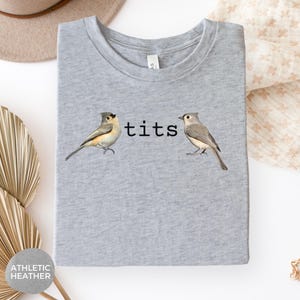 Puede incluir: Camiseta gris jaspeado con la palabra "tits" en texto negro entre dos ilustraciones de aves. Las aves est&aacute;n detalladas con tonos de gris, marr&oacute;n y blanco. La camiseta est&aacute; doblada y presentada sobre un fondo neutro.