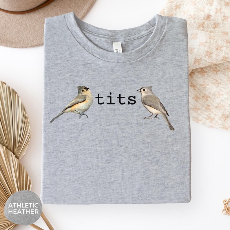 Puede incluir: Camiseta gris jaspeado con la palabra "tits" en texto negro entre dos ilustraciones de aves. Las aves est&aacute;n detalladas con tonos de gris, marr&oacute;n y blanco. La camiseta est&aacute; doblada y presentada sobre un fondo neutro.