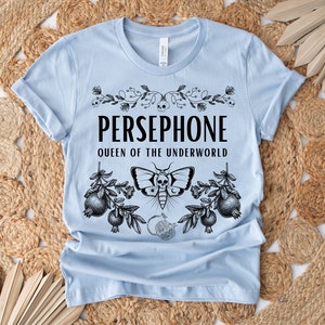 May include: Light blue t-shirt with a black graphic design featuring the text "PERSEPHONE QUEEN OF THE UNDERWORLD" with a floral design and a moth with a skull.