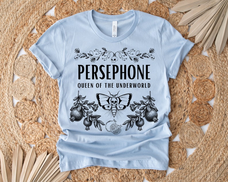 May include: Light blue t-shirt with a black graphic design featuring the text "PERSEPHONE QUEEN OF THE UNDERWORLD" with a floral design and a moth with a skull.