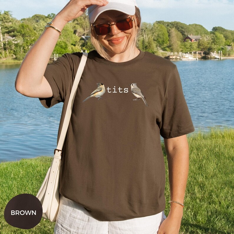 Puede incluir: Camiseta marr&oacute;n con la palabra "tits" en texto blanco y dos ilustraciones de p&aacute;jaros. La camiseta es de cuello redondo y manga corta. El fondo muestra una masa de agua y hierba verde.
