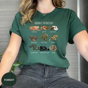 Puede incluir: Camiseta verde bosque con la frase "NOODLE ENTHUSIAST" y dibujos de serpientes como la serpiente del ma&iacute;z y la pit&oacute;n bola. La camiseta tiene mangas cortas y cuello redondo.