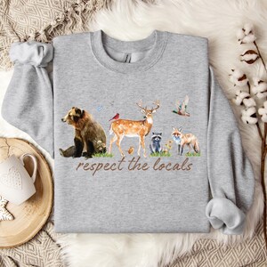 Op de afbeelding: Een grijze sweatshirt met een aquarel illustratie van bosdieren, waaronder een beer, een hert, een vos en een wasbeer. De zin "respect the locals" is in bruin schrift gedrukt. De sweatshirt heeft lange mouwen en een ronde hals.