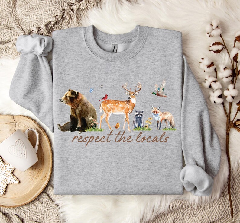 Op de afbeelding: Een grijze sweatshirt met een aquarel illustratie van bosdieren, waaronder een beer, een hert, een vos en een wasbeer. De zin "respect the locals" is in bruin schrift gedrukt. De sweatshirt heeft lange mouwen en een ronde hals.