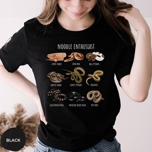 Puede incluir: Camiseta negra con la frase "NOODLE ENTHUSIAST" e ilustraciones de varias especies de serpientes, incluyendo serpiente del ma&iacute;z, boa de arena y pit&oacute;n bola. La camiseta tambi&eacute;n presenta culebra de liga, pit&oacute;n alfombra, culebra hocico de cerdo, rey de California, rey negro mexicano y boa rosada.