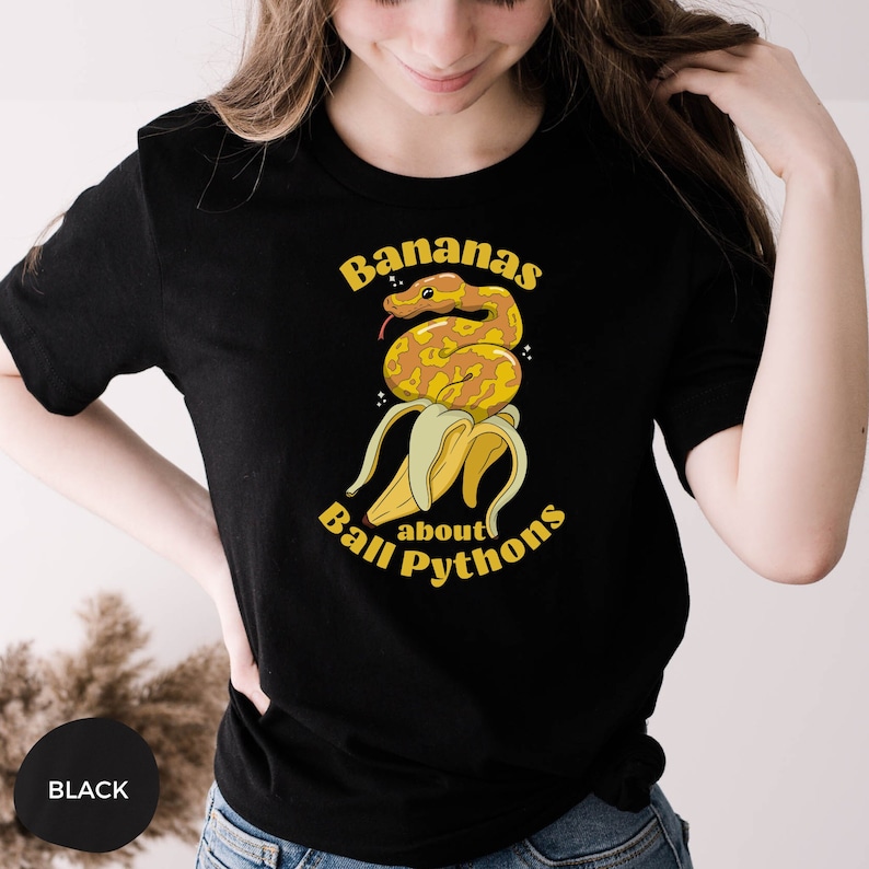 Op de afbeelding: Een zwart T-shirt met een gele banaan en een slang eromheen gewikkeld. De tekst "Bananas about Ball Pythons" staat op het T-shirt gedrukt.