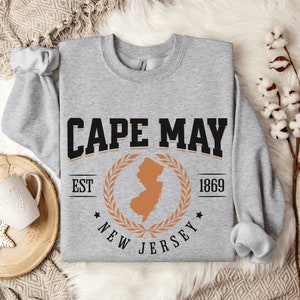 May include: Gray sweatshirt with a black and orange graphic design. The graphic features the text "CAPE MAY EST 1869 NEW JERSEY" with a laurel wreath and an outline of the state of New Jersey.