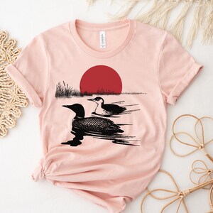 May include: A pink t-shirt with a black and white illustration of two loons swimming in a lake with a red sun setting in the background.