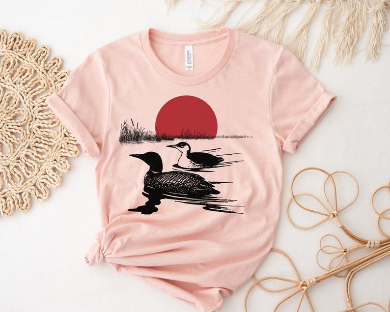 May include: A pink t-shirt with a black and white illustration of two loons swimming in a lake with a red sun setting in the background.