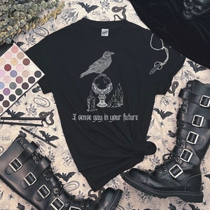 May include: Black t-shirt with a white graphic of a raven perched on a crystal ball with candles. The text "I sense gay in your future" is printed below the graphic.