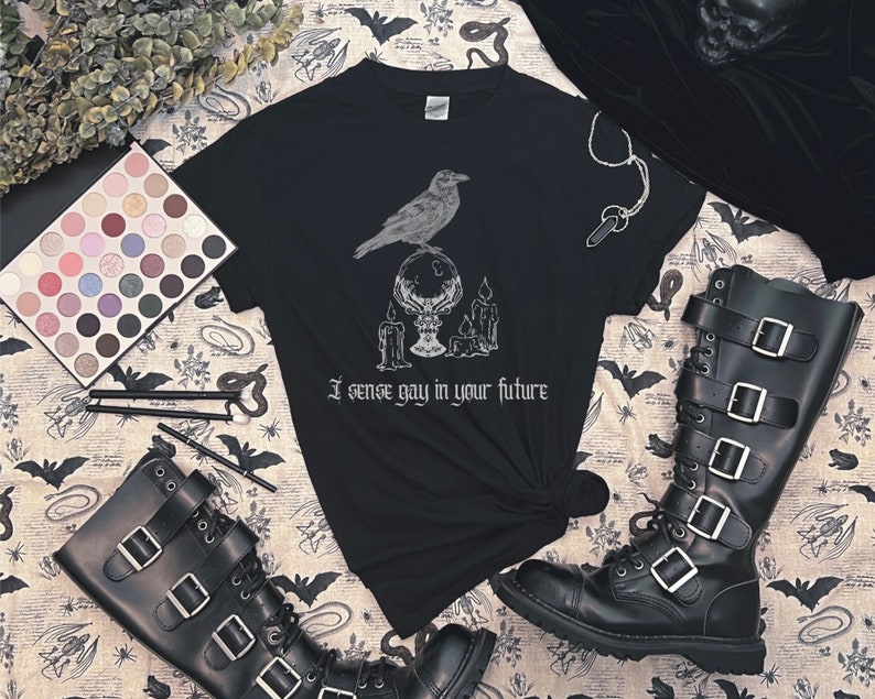 May include: Black t-shirt with a white graphic of a raven perched on a crystal ball with candles. The text "I sense gay in your future" is printed below the graphic.