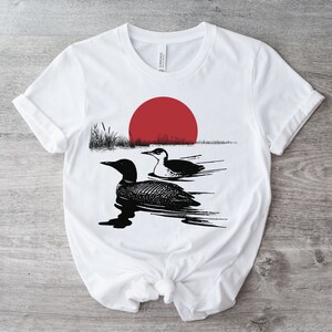 May include: White t-shirt with a black and white illustration of two loons swimming in a lake with a red sun setting in the background.