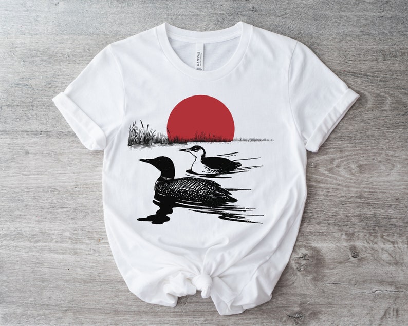 May include: White t-shirt with a black and white illustration of two loons swimming in a lake with a red sun setting in the background.