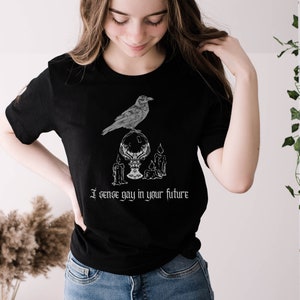 May include: A black t-shirt with a white graphic of a raven perched on a crystal ball with candles. The text "I sense gay in your future" is printed below the graphic.