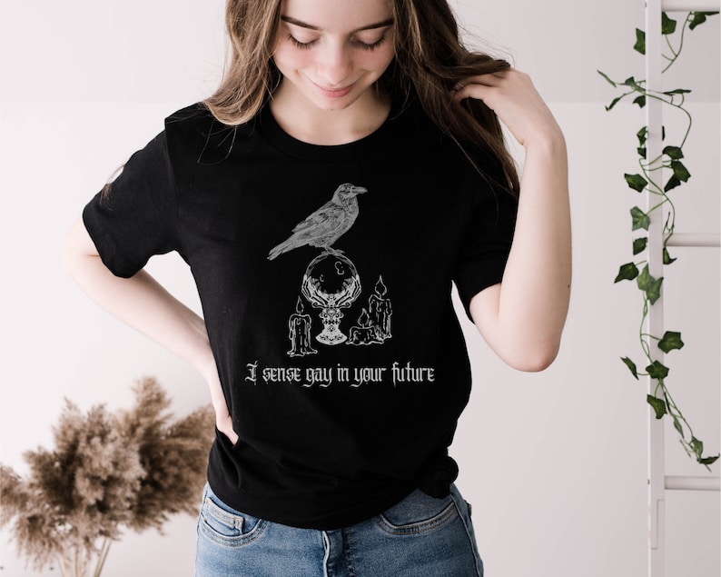 May include: A black t-shirt with a white graphic of a raven perched on a crystal ball with candles. The text "I sense gay in your future" is printed below the graphic.