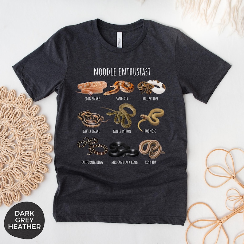 Puede incluir: Camiseta gris oscuro jaspeado con el texto "NOODLE ENTHUSIAST" e ilustraciones de varias especies de serpientes, incluyendo serpiente del ma&iacute;z, boa de arena y pit&oacute;n bola. La camiseta tambi&eacute;n presenta culebra de liga, pit&oacute;n alfombra y culebra hocico de cerdo.