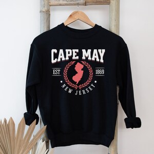 May include: Black sweatshirt with a white graphic print of a red outline of the state of New Jersey with a laurel wreath around it. The text "CAPE MAY EST 1869 NEW JERSEY" is printed on the sweatshirt.