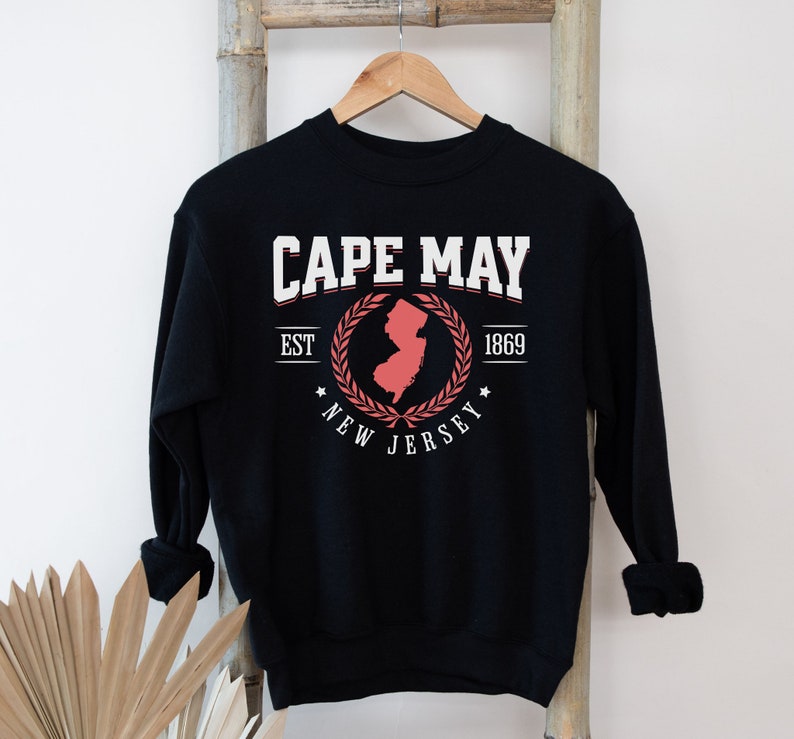 May include: Black sweatshirt with a white graphic print of a red outline of the state of New Jersey with a laurel wreath around it. The text "CAPE MAY EST 1869 NEW JERSEY" is printed on the sweatshirt.