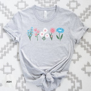 May include: A gray t-shirt with a row of five colorful flowers printed on the front. The flowers are blue, pink, white, and green.